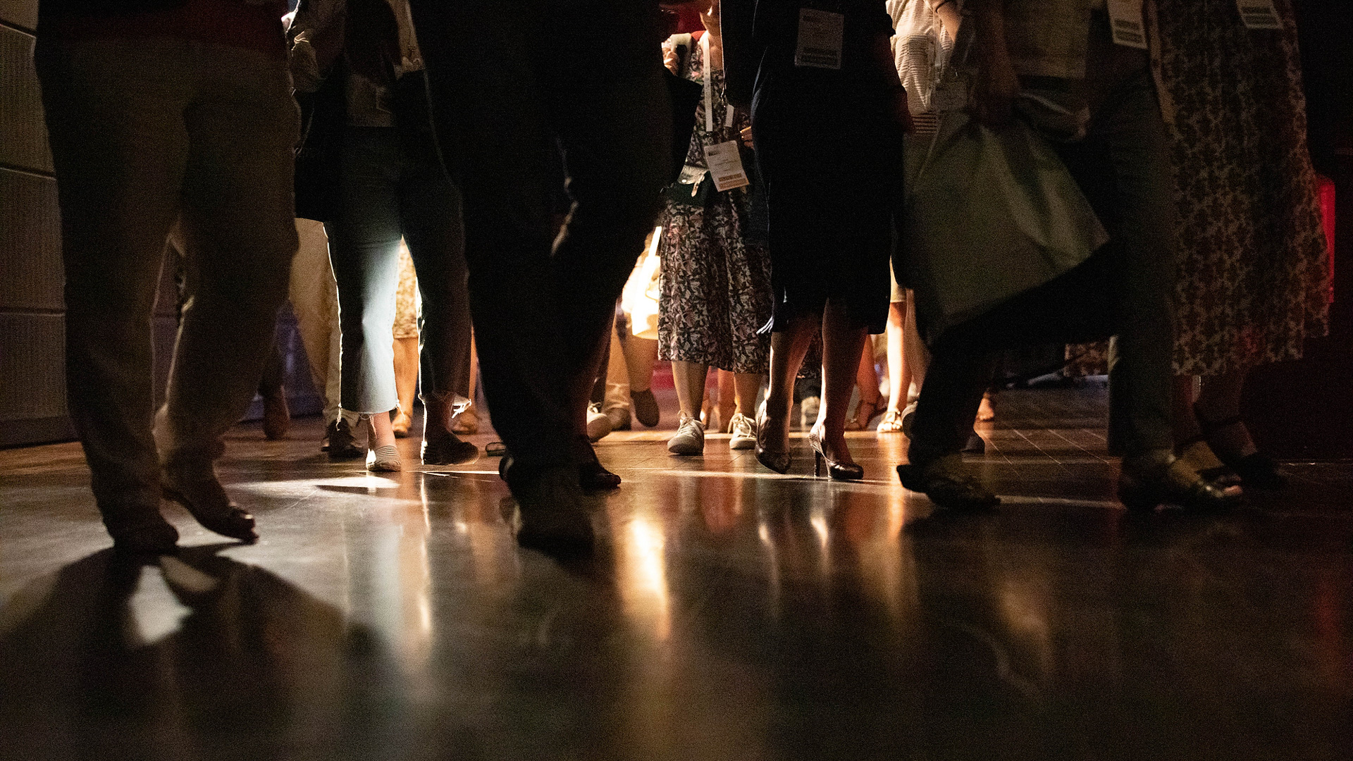 Gros plan sur les jambes et les pieds de plusieurs personnes marchant ou debout sur un sol brillant, reflétant les lumières du lieu. La scène est sombre avec des reflets lumineux.