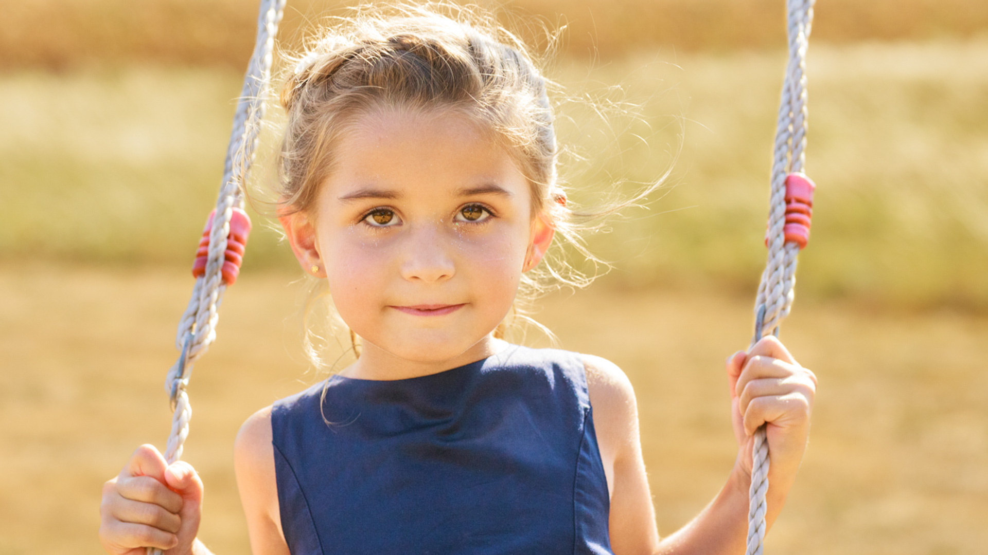 Gros plan d'une jeune fille aux cheveux châtains clairs tressés, vêtue d'une robe bleu foncé, regardant directement l'appareil photo tout en tenant les cordes d'une balançoire.