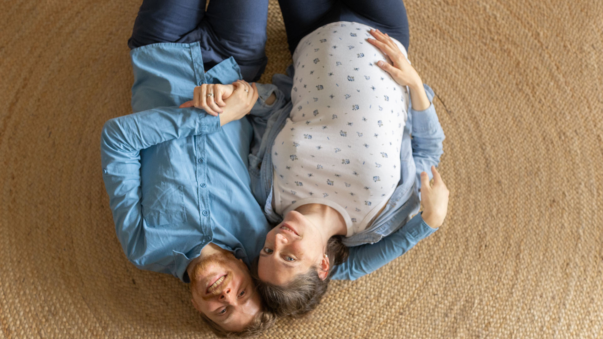 Vue de dessus d'un couple, la femme visiblement enceinte, allongés sur un tapis tissé, se tenant la main et souriant à l'appareil photo.