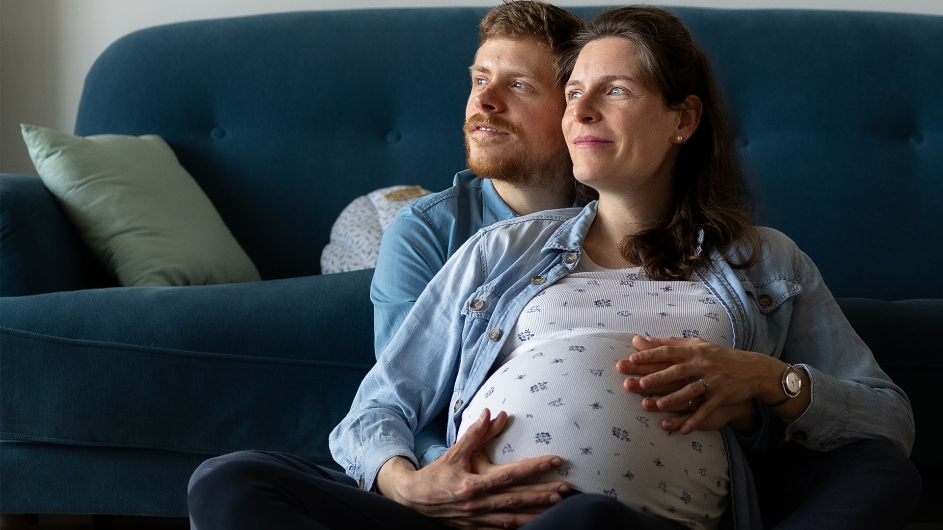 Une femme enceinte et un homme assis ensemble par terre devant un canapé bleu. L'homme a son bras autour de la femme, qui tient son ventre de femme enceinte, tous deux regardant sur le côté.