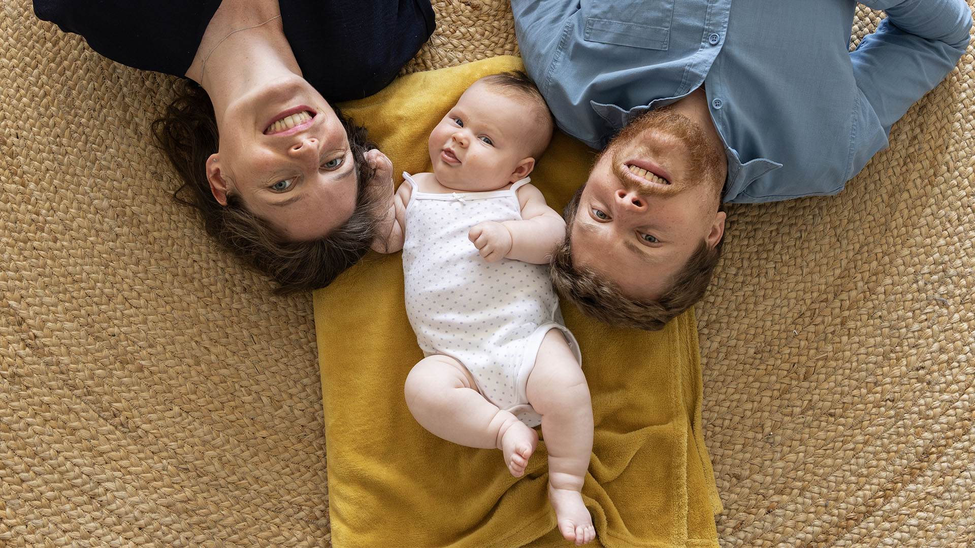 Vue de dessus d'une famille (mère, père et un bébé) allongée sur une couverture jaune sur un tapis tissé, regardant l'appareil photo et souriant. Le bébé est au centre.