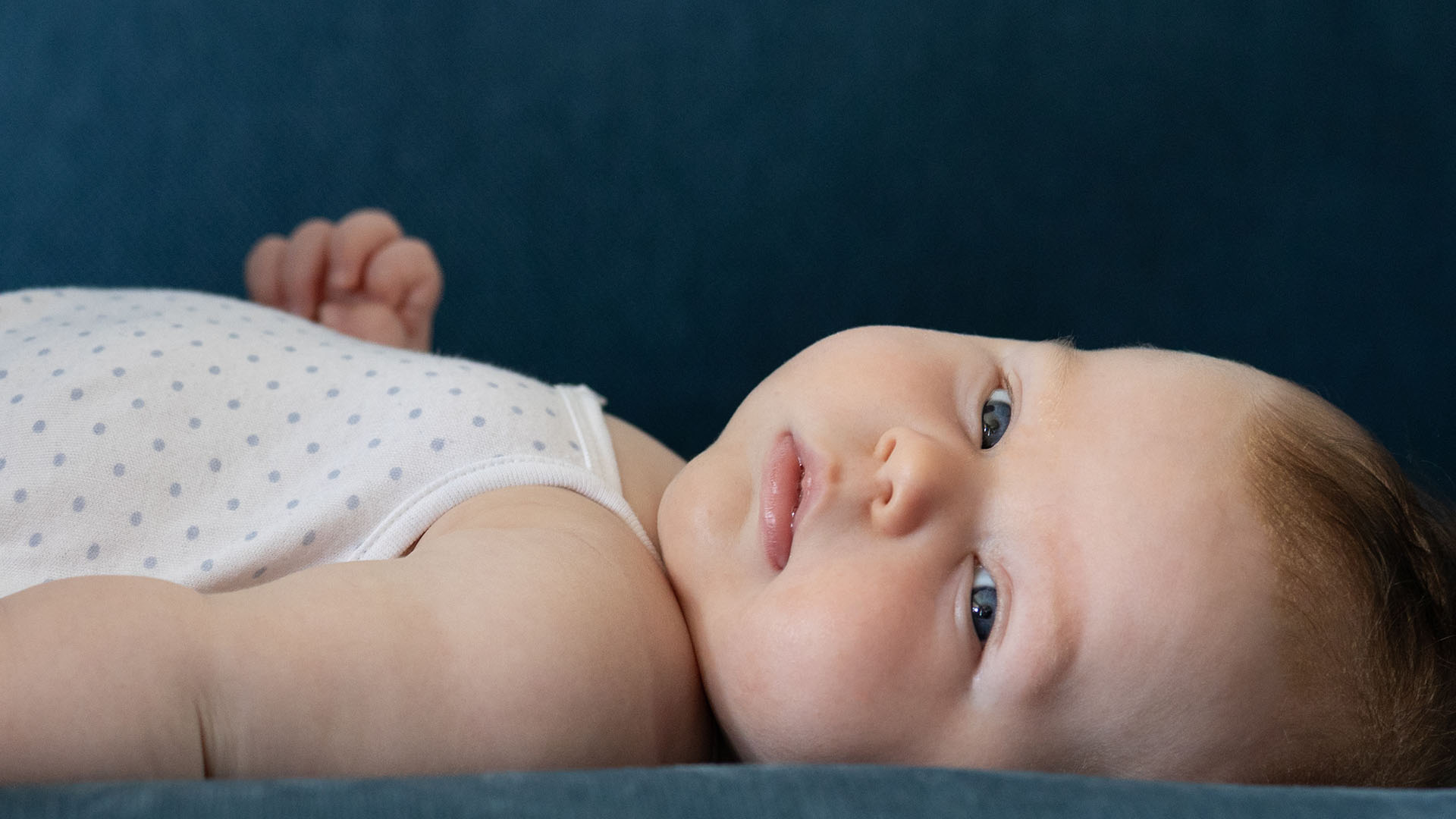 Gros plan d'un bébé allongé sur une surface bleue, regardant directement l'appareil photo avec de grands yeux bleus.