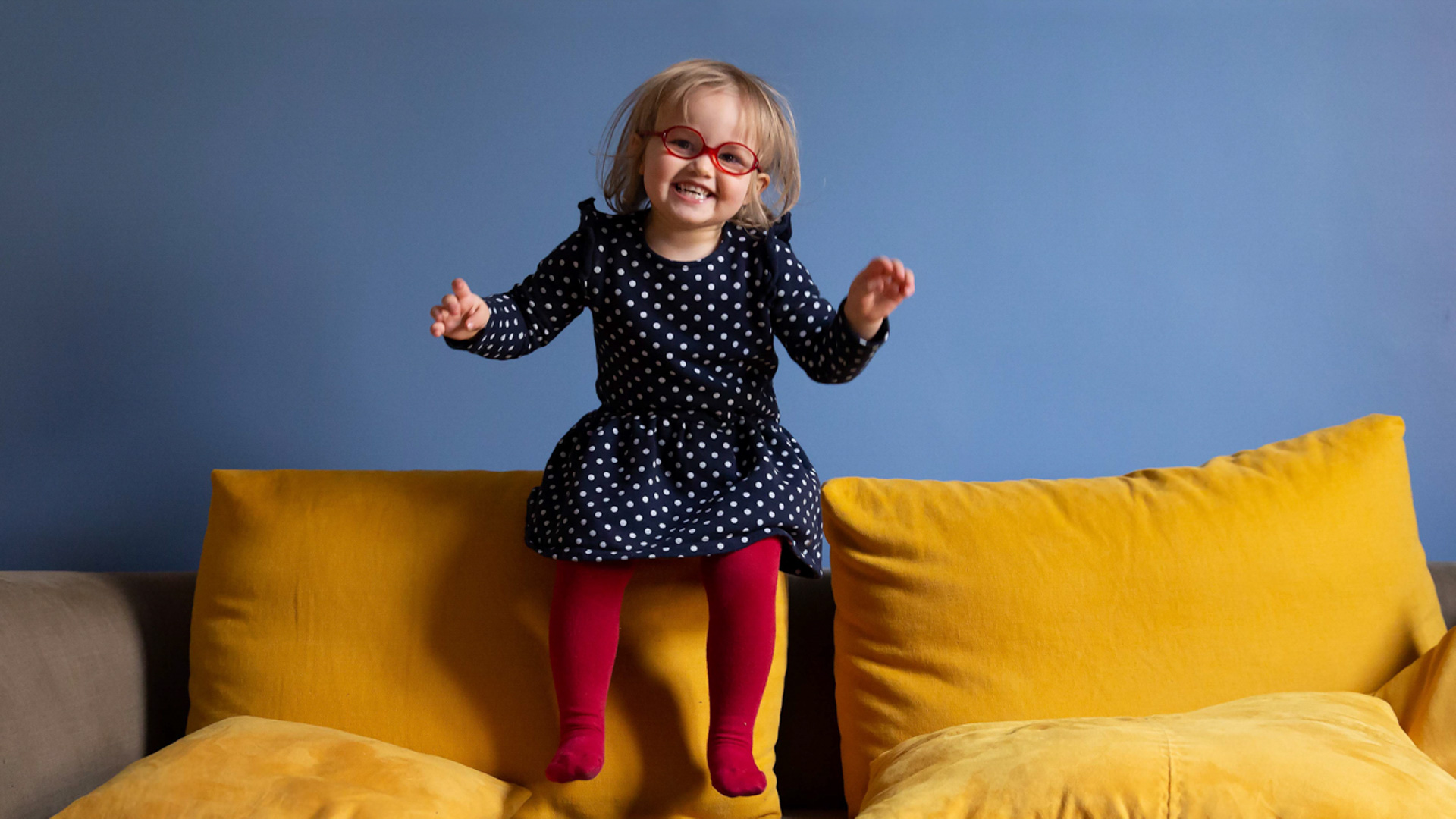 Une jeune fille aux cheveux blonds et aux lunettes rouges, vêtue d'une robe à pois et de collants rouges, sautant joyeusement sur des coussins de canapé jaunes devant un mur bleu.
