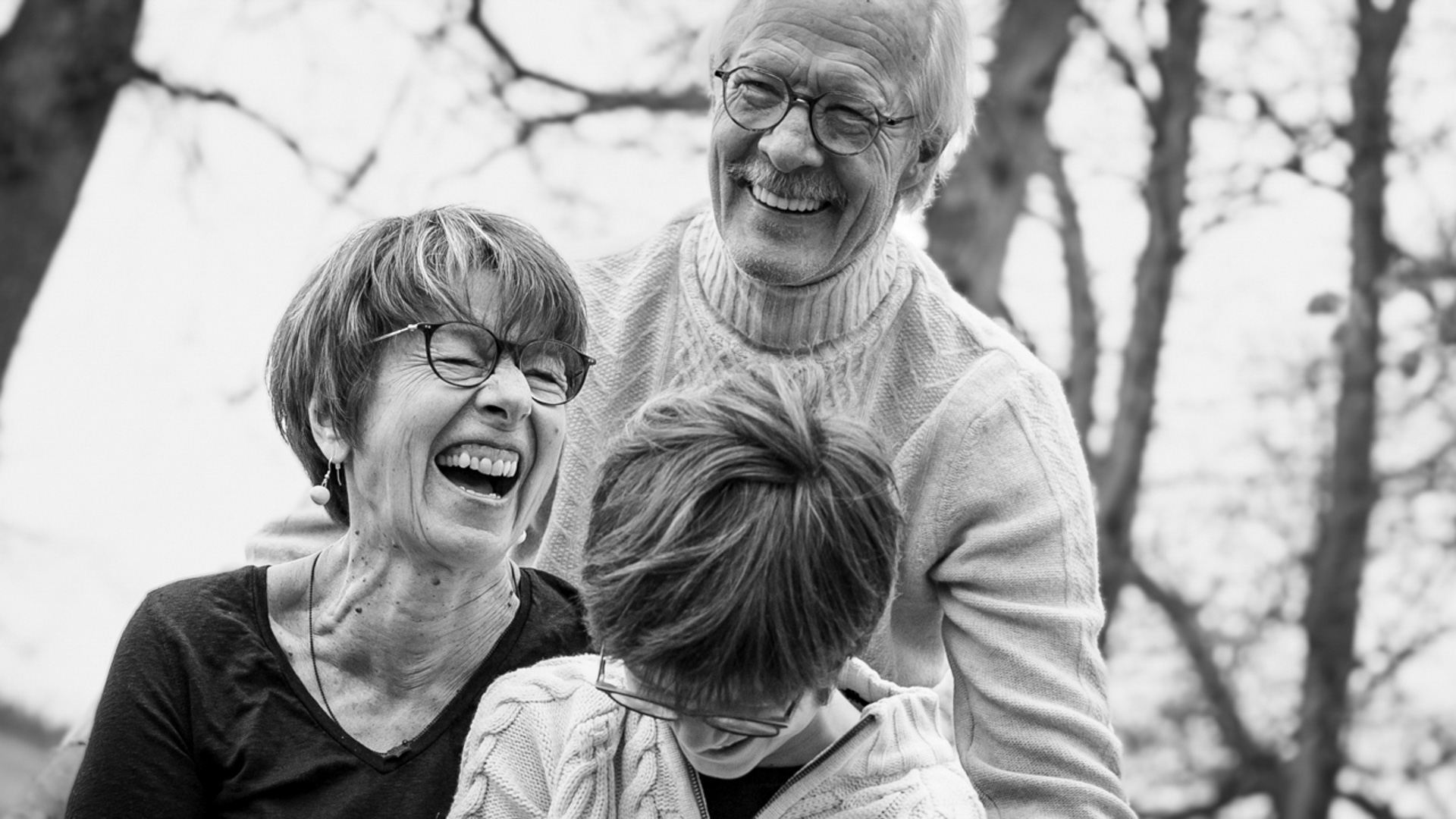 Photo en noir et blanc d'un couple âgé et d'un enfant, tous riant. L'homme est derrière, et la femme et l'enfant sont devant, regardant vers le bas et souriant.