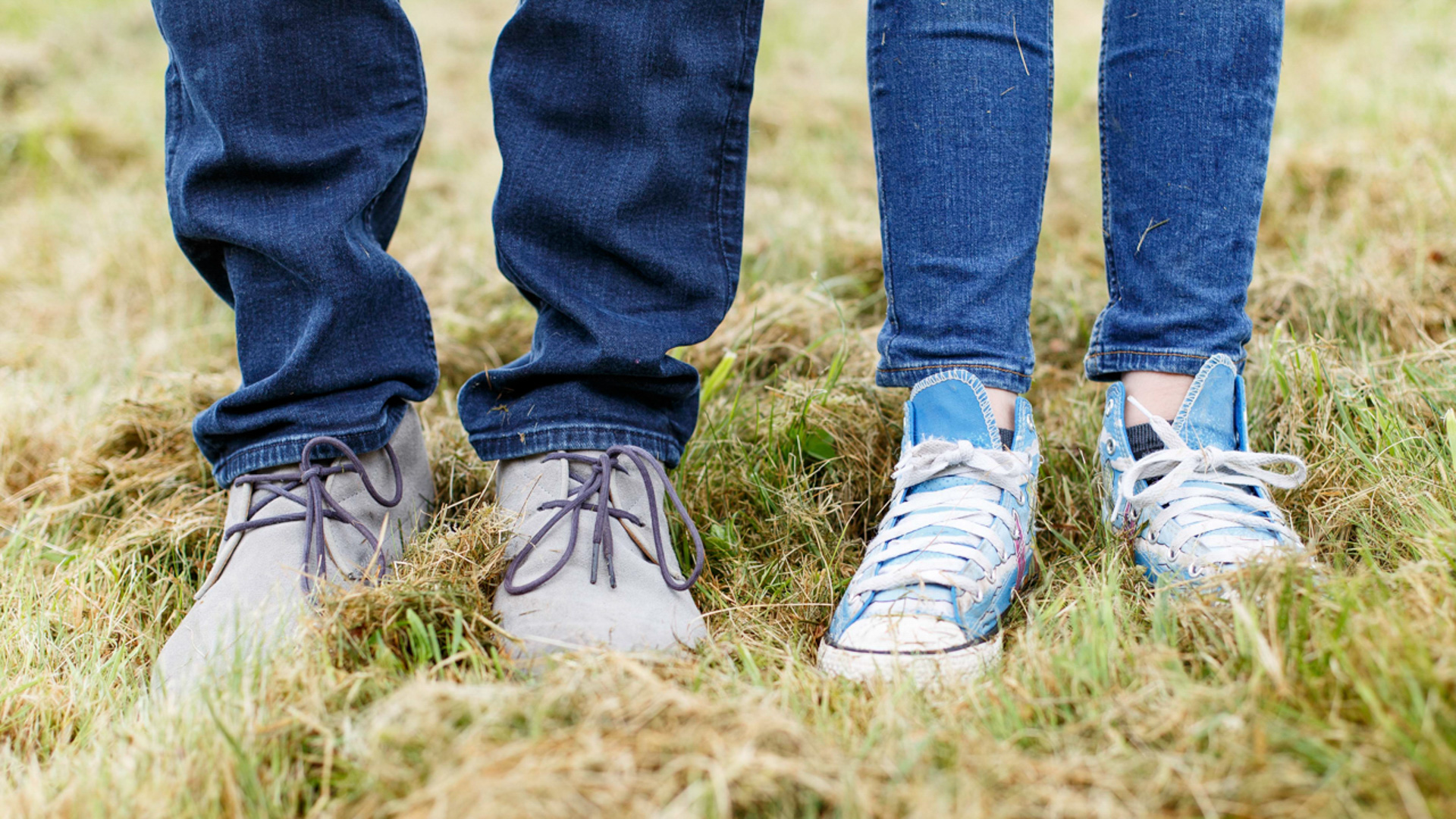 Gros plan de deux paires de jambes vêtues de jeans et de baskets, debout dans de l'herbe haute verte et brune. Une paire porte des chaussures grises, l'autre des Converse bleues et blanches.