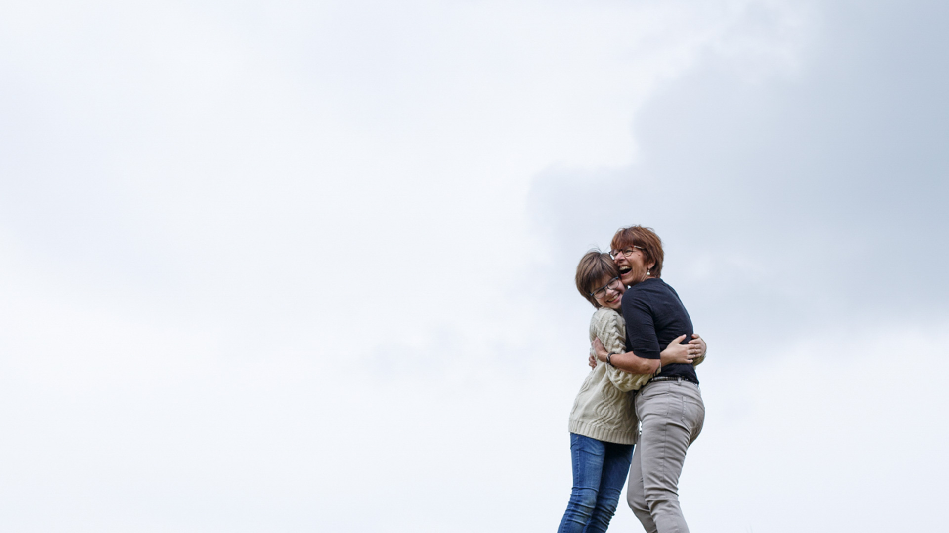 Deux femmes, l'une aux cheveux courts et portant des lunettes, l'autre aux cheveux plus longs et portant également des lunettes, s'embrassant et riant en extérieur sous un ciel clair et nuageux.