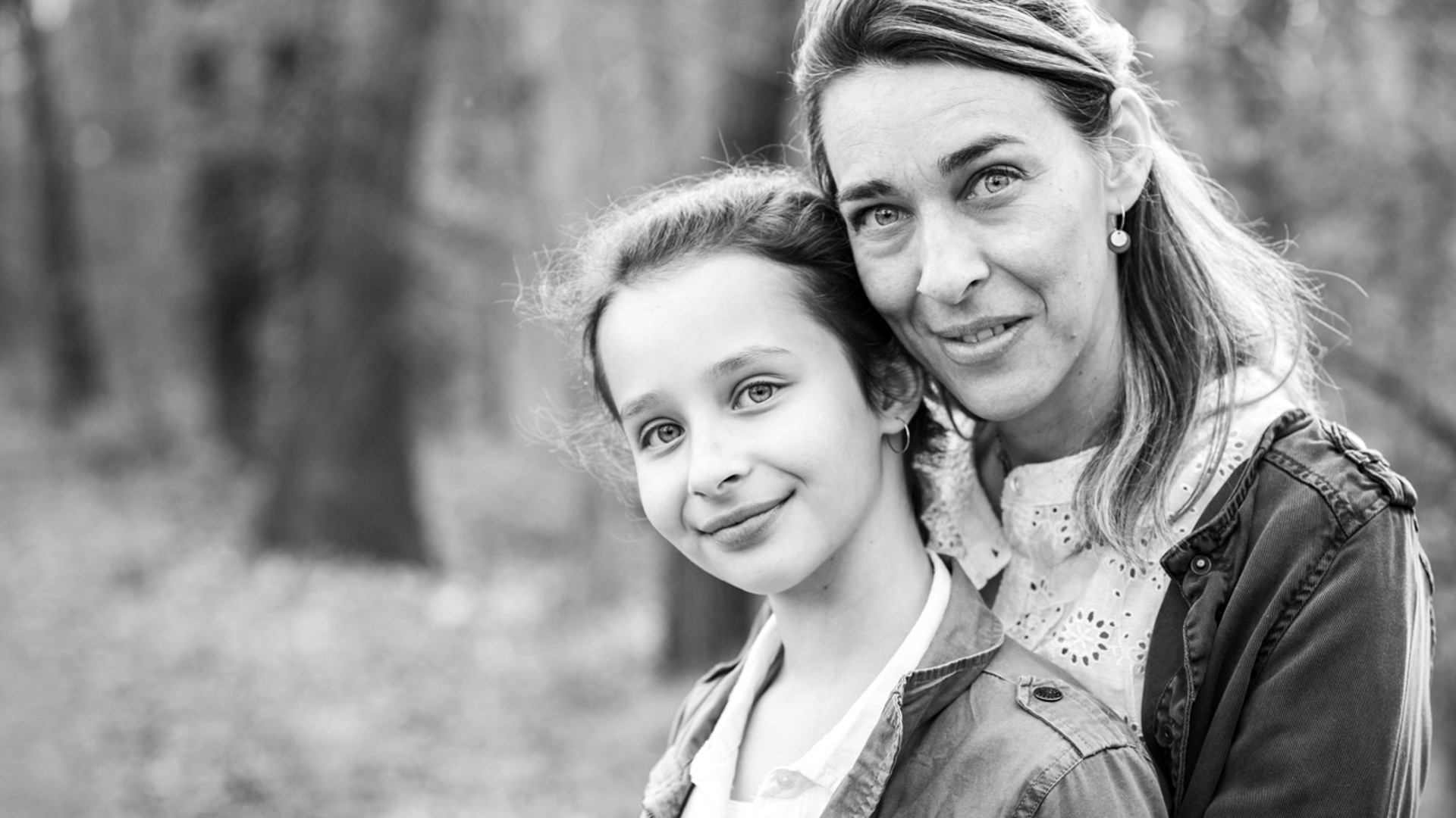 Gros plan en noir et blanc d'une mère et de sa fille. La mère est derrière, regardant directement l'appareil photo avec son bras autour de la fille, qui regarde également l'appareil photo et sourit doucement.