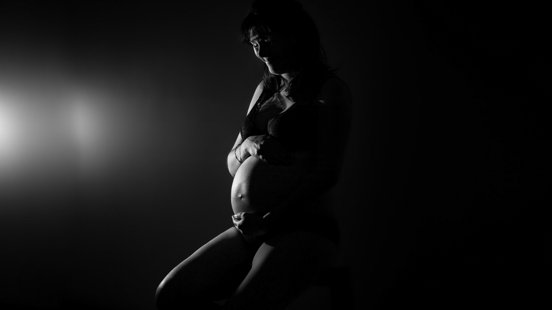 Silhouette en noir et blanc d'une femme enceinte en lingerie, assise et tenant doucement son ventre, avec une source de lumière dramatique venant de la gauche.