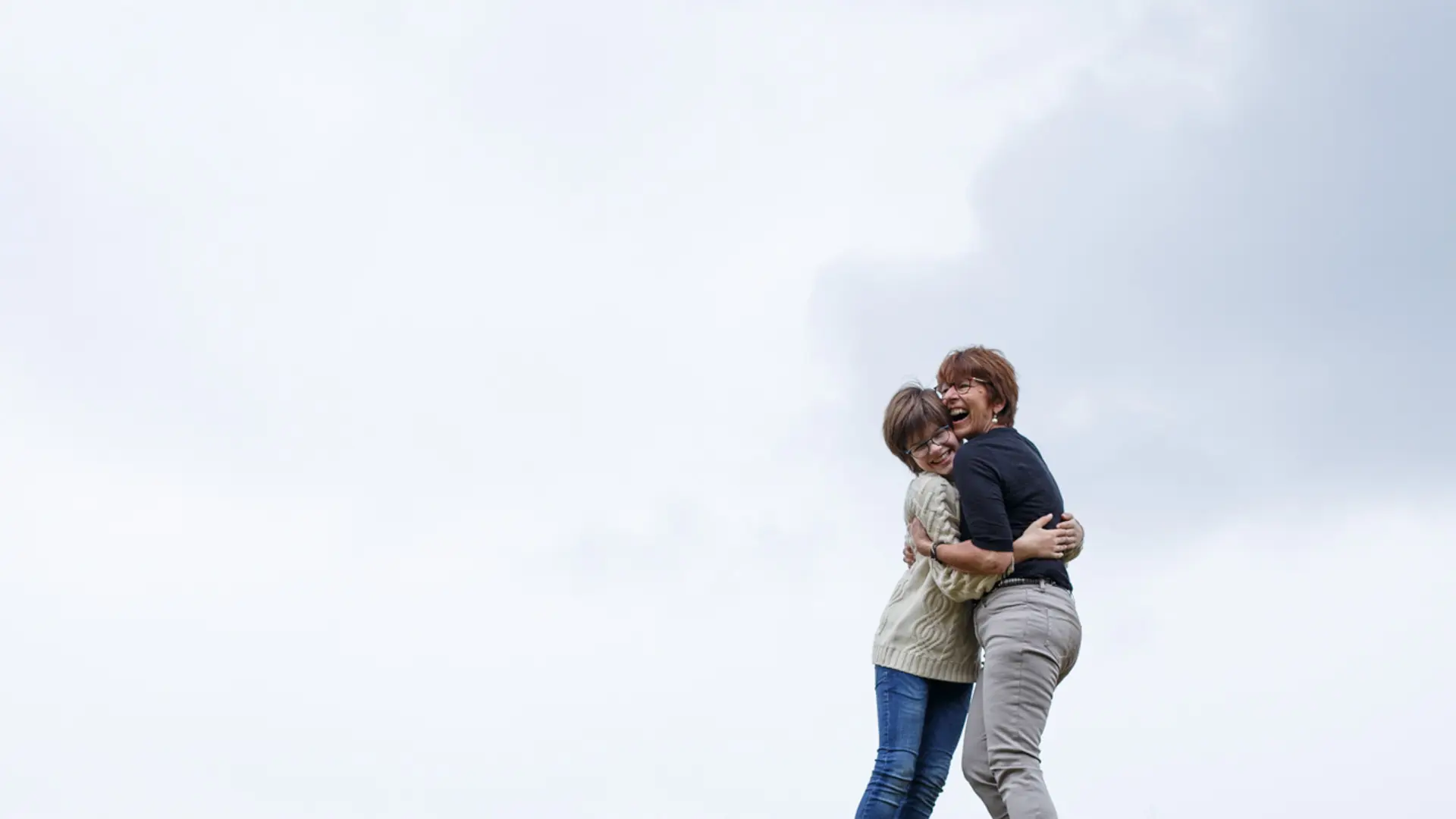 Une mariée en robe blanche légère est lancée en l'air par un groupe d'hommes. Ses bras sont levés et elle semble rire, sur un fond de ciel bleu avec des nuages et des collines.