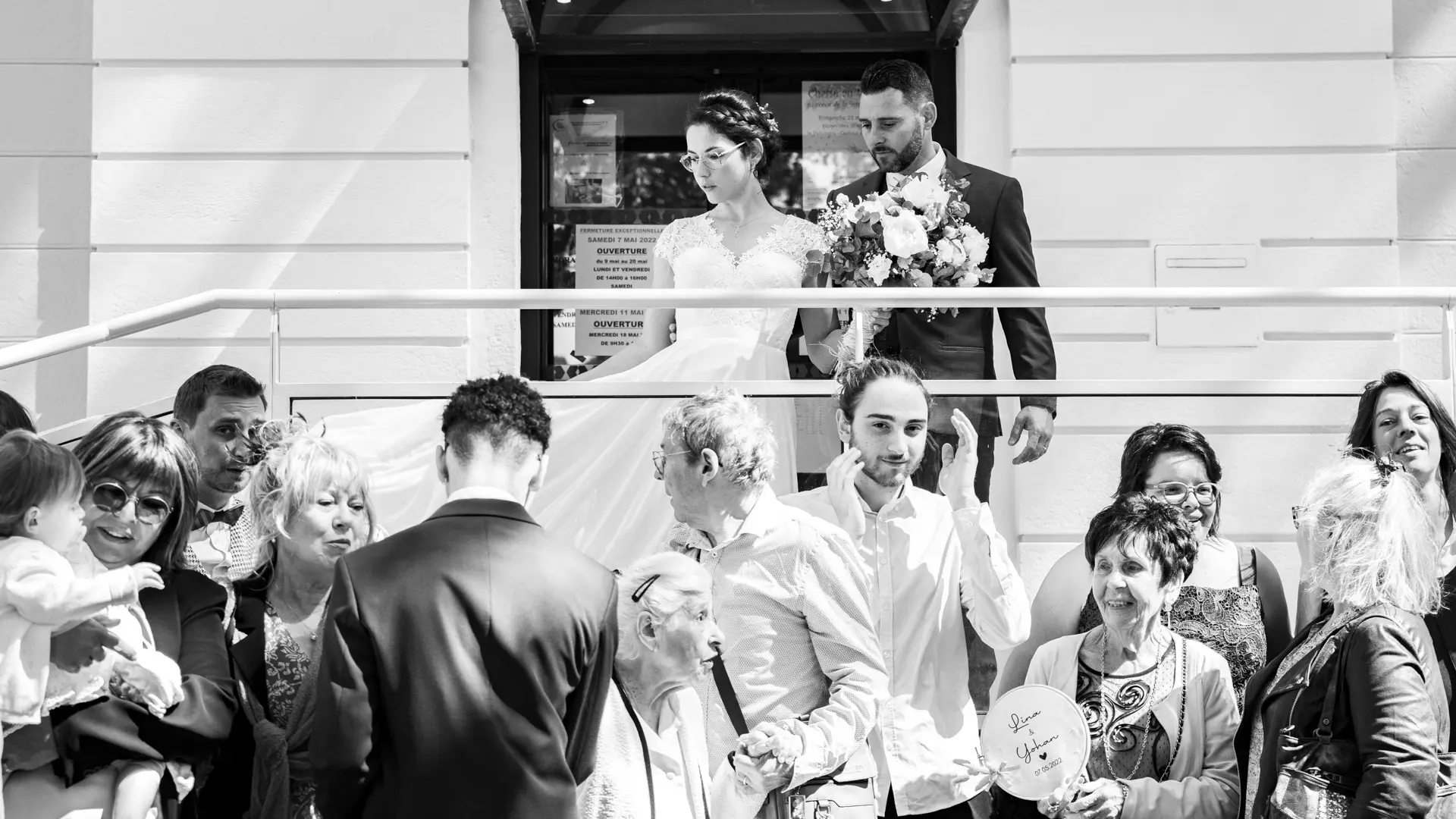 Photo en noir et blanc d'un couple de mariés s'embrassant tendrement devant une porte vitrée, avec des bulles de savon flottant autour d'eux. Un homme âgé et d'autres invités souriants sont visibles en arrière-plan.