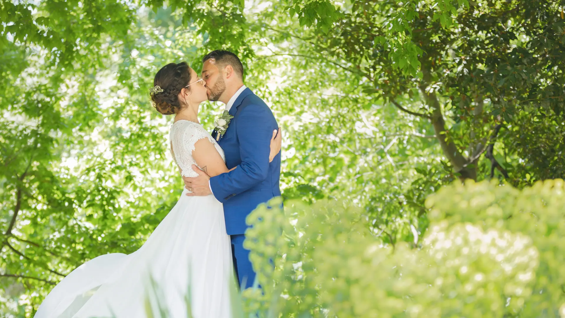 Un groupe d'hommes en chemises claires lancent une mariée en robe blanche en l'air. La mariée rit en étant soulevée, tandis que les hommes en dessous la rattrapent, sur un fond de ciel bleu et de paysage vallonné.