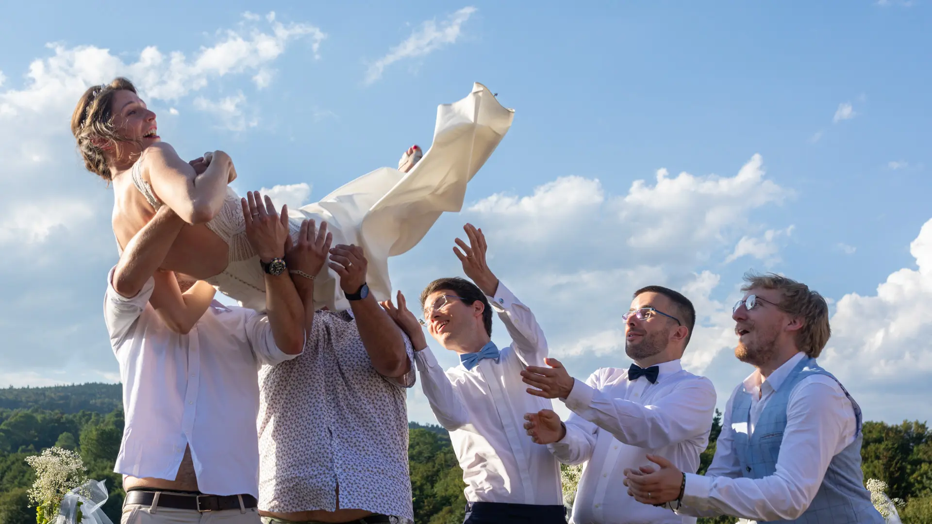 Une mariée en robe blanche légère est lancée en l'air par un groupe d'hommes, vus de dos. Elle a les bras levés et semble rire de joie, sur un fond de ciel bleu avec des nuages et des collines verdoyantes.
