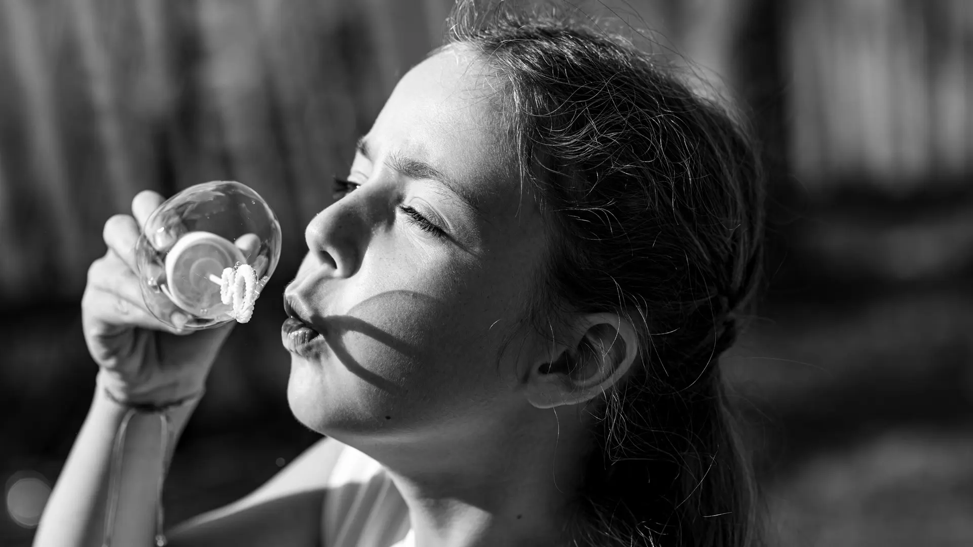 Photo en noir et blanc d'une jeune fille soufflant des bulles, les yeux fermés et le visage concentré, avec le soleil créant des ombres sur sa joue. Elle tient un petit récipient à bulles près de sa bouche.